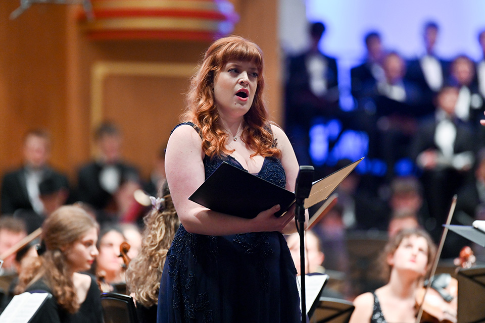 A soprano, wearing a formal navy dress, performing in an orchestral performance, with an orchestra and choir behind her.